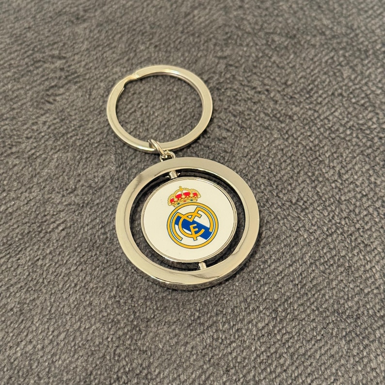 May include: A silver keyring with a circular pendant displaying the Real Madrid logo. The logo incorporates a crown, a blue and yellow design, and the team's initials. The pendant rotates and is attached to a silver key ring.