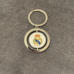 May include: Silver keyring with a circular pendant displaying the Real Madrid crest. The crest features a crown, a circular emblem with blue and yellow details, and the letters "RMCF". The keyring has a silver ring for attaching keys.