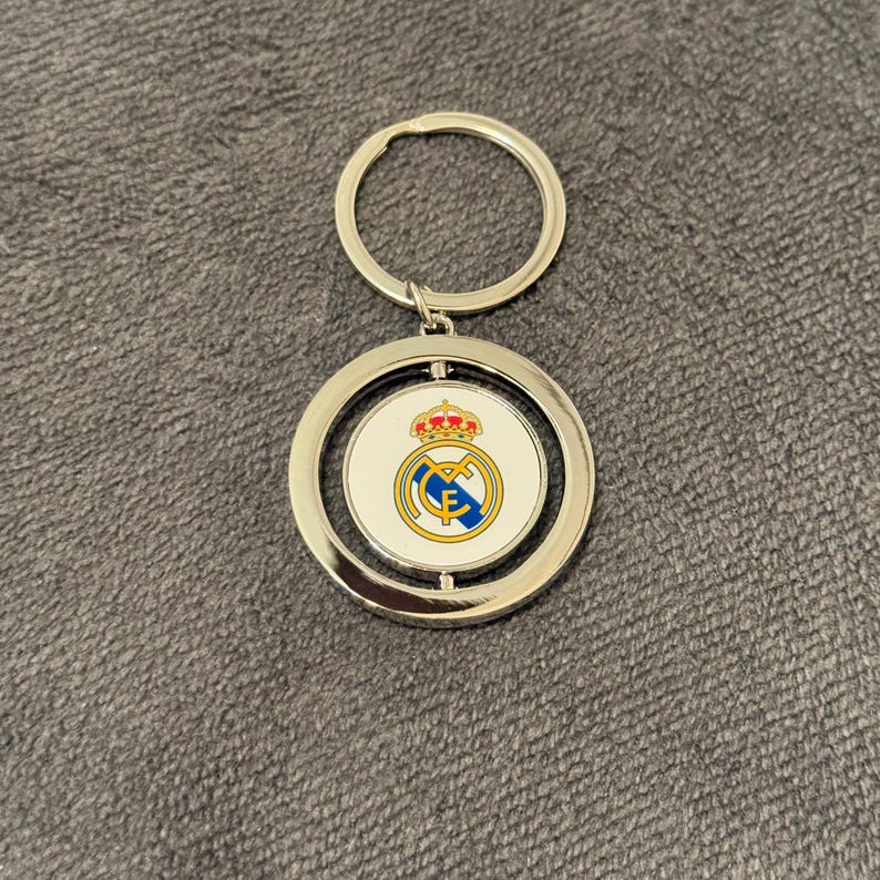 May include: Silver keyring with a circular pendant displaying the Real Madrid crest. The crest features a crown, a circular emblem with blue and yellow details, and the letters "RMCF". The keyring has a silver ring for attaching keys.
