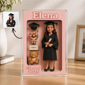 May include: A graduation-themed display case with the name "Elena" in pink lettering. Inside, a figurine in a graduation gown holds a diploma. Additional elements include a graduation cap, flowers, a diploma, and a teddy bear. The text "Class of 2025" is also visible.