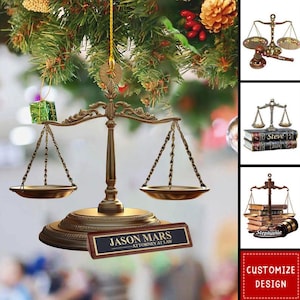 May include: A gold-colored scale of justice ornament with a nameplate that reads "JASON MARS, ATTORNEY AT LAW." The ornament is hanging from a Christmas tree with green needles, a small gift, and other decorations.