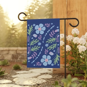 May include: A blue garden flag with a floral pattern featuring blue, pink, and green flowers and leaves. The flag is hanging on a black metal stand.