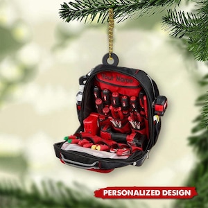 Puede incluir: Adorno navideño de bolsa de herramientas negra y roja con una cadena dorada. La bolsa está abierta, revelando varias herramientas rojas. El texto "Your Name" está encima de la abertura de la bolsa y "PERSONALIZED DESIGN" en la parte inferior.