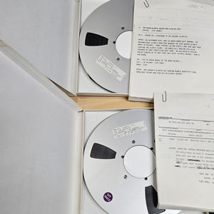 May include: Two open white boxes containing large silver audio recording reels. Each reel has a black tape and a white label. Beside the reels are several sheets of paper with typed text and a paper clip.