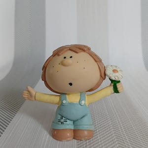 Könnte beinhalten: Figur einer Zeichentrickfigur mit gelbem Oberteil und blauer Latzhose. Die Figur hat braunes Haar, Sommersprossen und hält eine weiße Gänseblümchen mit grünem Stiel. Die Arme der Figur sind ausgestreckt.
