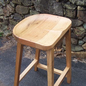 May include: A light-colored wooden stool with a curved seat and four sturdy legs. The stool is made of natural wood, showcasing the grain and texture. The design is simple and functional, suitable for various settings.