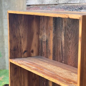 May include: A rustic, wooden bookshelf with two shelves. The wood has a natural, warm brown tone with visible grain and knots, giving it a reclaimed or vintage appearance. The shelves are sturdy and ready for display.
