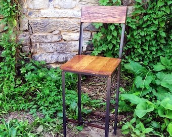 Puede incluir: Un taburete de bar de madera y metal con un asiento de madera marrón y un marco de metal negro. El taburete está sentado en un camino de tierra con hierba verde y plantas en el fondo.