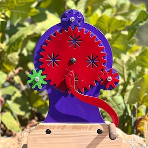 May include: A purple and red wooden gear toy with a crank handle. The toy has a base made of wood and a purple gear mechanism with red gears. The gears are decorated with a star pattern.