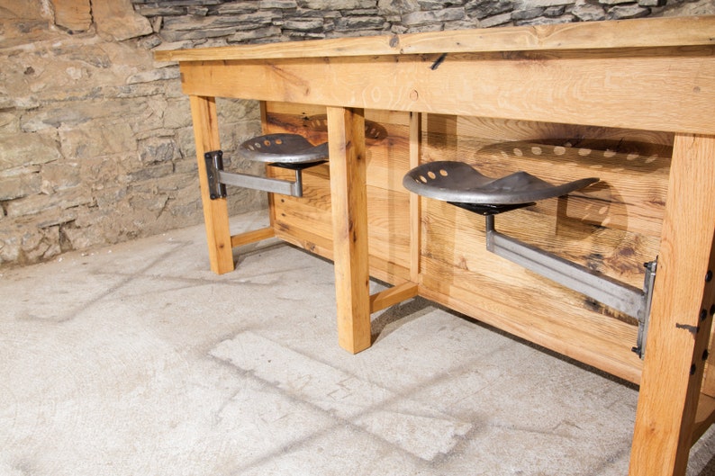 May include: A rustic wooden table with two metal industrial-style swivel seats attached to the underside. The table is made of reclaimed wood and has a natural finish. The seats are made of metal and have a black finish.