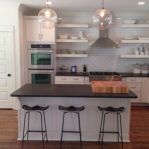 May include: A white kitchen with black countertops and white cabinets. The kitchen has a large island with three bar stools. The island has a wooden cutting board on top. The kitchen also has a stainless steel range hood and double ovens.