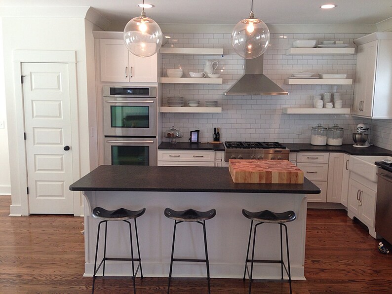 May include: A white kitchen with black countertops and white cabinets. The kitchen has a large island with three bar stools. The island has a wooden cutting board on top. The kitchen also has a stainless steel range hood and double ovens.