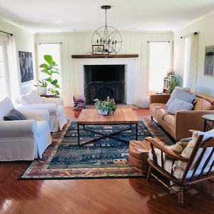 May include: A living room with a fireplace, a brown leather sofa, two white armchairs, a coffee table, a rug, and a wooden rocking chair. The fireplace has a black wood-burning stove and a white brick surround. The rug is blue and green with a floral pattern. The coffee table is made of wood and has a black metal frame.