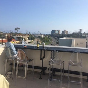 May include: A rooftop patio with a black metal bar table and four clear plastic chairs. A person is sitting at the table with two wine bottles and glasses.