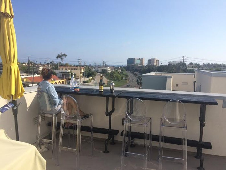 May include: A rooftop patio with a black metal bar table and four clear plastic chairs. A person is sitting at the table with two wine bottles and glasses.