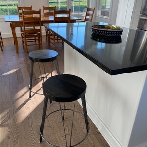 May include: Two black metal and wood bar stools with a black countertop in the background. The stools have a round seat and a wire frame.