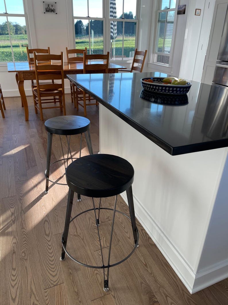 May include: Two black metal and wood bar stools with a black countertop in the background. The stools have a round seat and a wire frame.