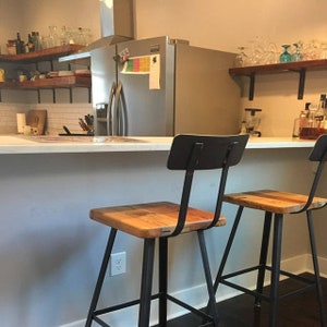May include: Two rustic bar stools with square wooden seats and black metal frames. The stools have a simple design with a backrest and are positioned in front of a kitchen counter. The kitchen has wooden shelves and a stainless steel fridge.