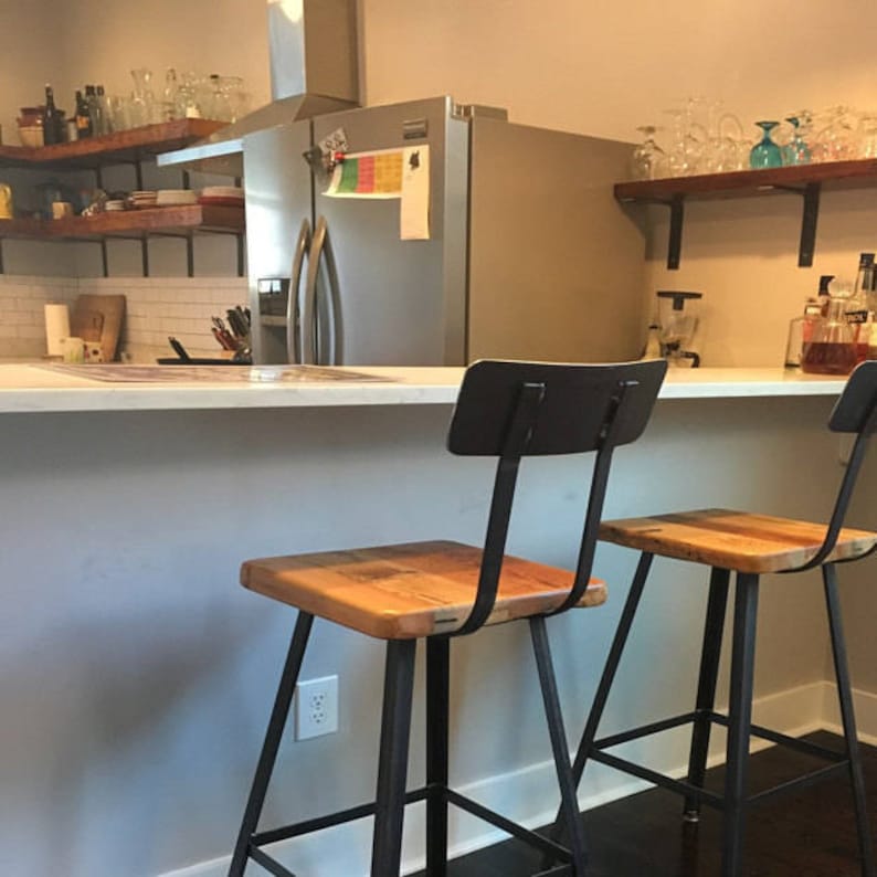 May include: Two rustic bar stools with square wooden seats and black metal frames. The stools have a simple design with a backrest and are positioned in front of a kitchen counter. The kitchen has wooden shelves and a stainless steel fridge.