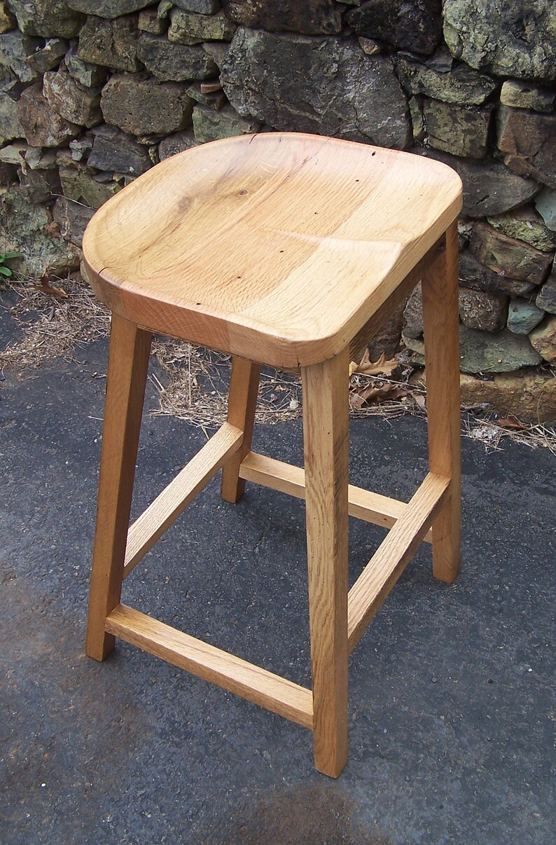 May include: A light-colored wooden stool with a curved seat and four sturdy legs. The stool is made of natural wood, showcasing the grain and texture. The design is simple and functional, suitable for various settings.