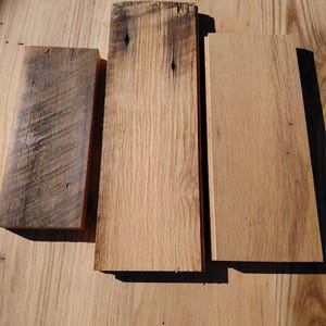May include: Three rectangular wooden planks of varying shades of brown and tan are arranged on a light brown wooden surface. The planks show natural wood grain and knots, with one plank appearing weathered and darker in color.