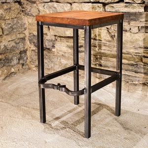May include: A handcrafted bar stool with a square, solid wood seat and a dark metal frame. The seat is a warm brown color, and the metal frame has a raw, industrial finish. The stool has a footrest and is suitable for a kitchen or bar setting.