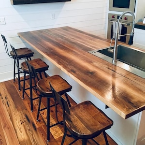 May include: A kitchen island with a wooden countertop and a stainless steel sink. Four wooden bar stools with black metal legs are lined up in front of the island. A shelf on the wall holds a clock, a horseshoe, and a red bull figurine.