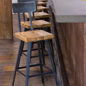 May include: A series of bar stools with black metal frames and wooden seats. The seats are square and made of light-coloured wood. The stools are arranged along a concrete bar, with a dark metal footrest.
