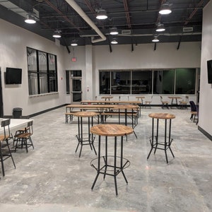 May include: An interior view of a modern, open-plan space, possibly a brewery or taproom. The room has a polished concrete floor, high ceilings with track lighting, and a variety of seating, including high tables with metal frames and wooden tops, and long communal tables.