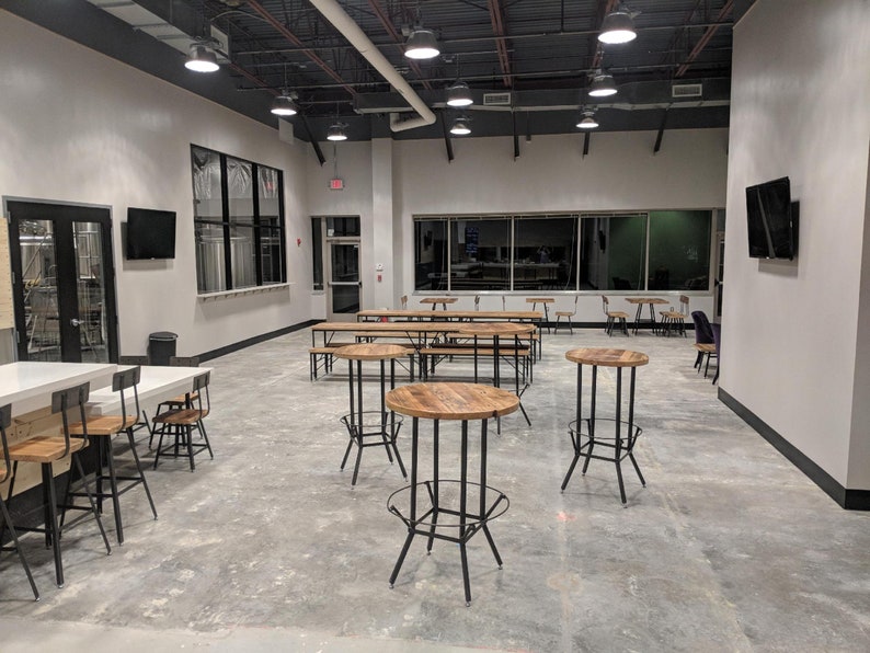 May include: An interior view of a modern, open-plan space, possibly a brewery or taproom. The room has a polished concrete floor, high ceilings with track lighting, and a variety of seating, including high tables with metal frames and wooden tops, and long communal tables.