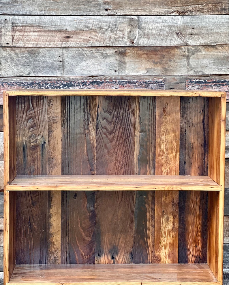May include: A rustic, two-tiered wooden shelf with a natural finish. The shelf is constructed from vertical planks of varying shades of brown and tan, creating a textured appearance. The frame is a lighter shade of wood, and the shelf is designed for storage or display.
