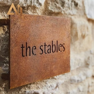 May include: A rectangular metal sign with a rusted, textured brown finish. The words "the stables" are cut out from the centre of the metal plate in a simple, lowercase sans-serif font, allowing the light to pass through the letters.