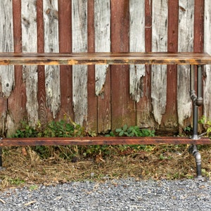 May include: A rustic wooden table with a metal pipe frame. The table has two shelves and is made of reclaimed wood. The table is in front of a weathered wooden fence.