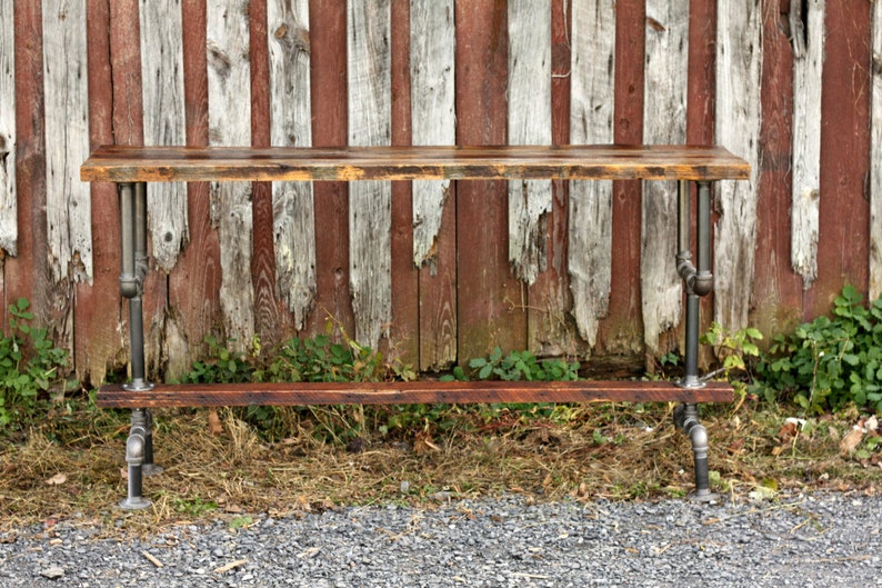 May include: A rustic wooden table with a metal pipe frame. The table has two shelves and is made of reclaimed wood. The table is in front of a weathered wooden fence.