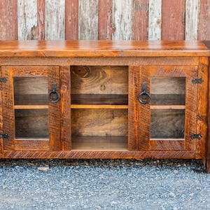 May include: A rustic wooden TV stand with three glass doors and two shelves. The stand has a dark brown stain and black metal hardware.