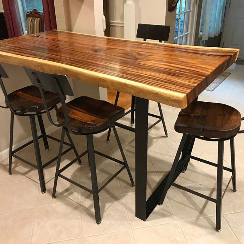 Op de afbeelding: Een houten tafel met een live edge blad en zwarte metalen poten. De tafel wordt omringd door vier zwarte metalen barkrukken met houten zittingen.