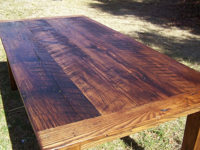 May include: A rustic wooden table with a dark brown stain. The table top is made of reclaimed wood planks with visible knots and grain patterns. The table has a simple, rectangular design with a slightly distressed finish.