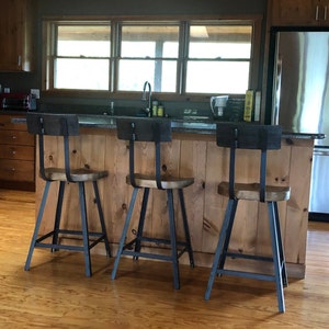 Op de afbeelding: Drie zwarte metalen barstoelen met houten zittingen en rugleuningen. De krukken staan voor een houten bar met een granieten werkblad. De krukken bevinden zich in een keuken met een koelkast van roestvrij staal en houten kasten.