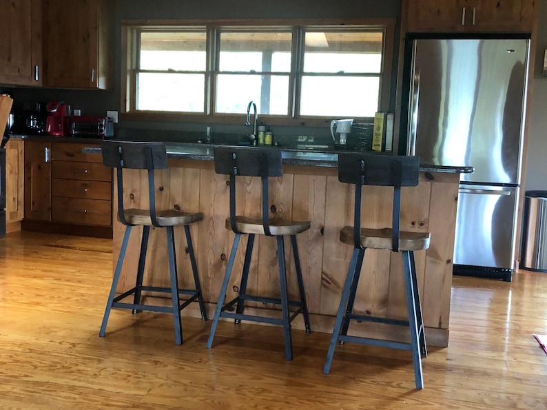 Op de afbeelding: Drie zwarte metalen barstoelen met houten zittingen en rugleuningen. De krukken staan voor een houten bar met een granieten werkblad. De krukken bevinden zich in een keuken met een koelkast van roestvrij staal en houten kasten.