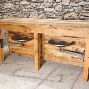 May include: A rustic wooden table with two metal tractor seat stools attached to the sides. The table is made of reclaimed wood and has a natural finish. The stools are black and have a simple design.