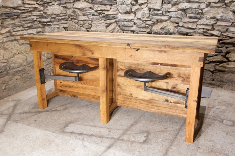 May include: A rustic wooden table with two metal tractor seat stools attached to the sides. The table is made of reclaimed wood and has a natural finish. The stools are black and have a simple design.