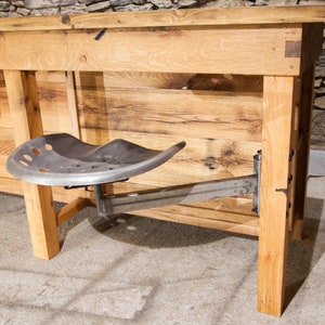 May include: A rustic wooden table with a metal tractor seat attached to the side. The table is made of reclaimed wood and has a natural finish. The seat is made of metal and has a black finish.