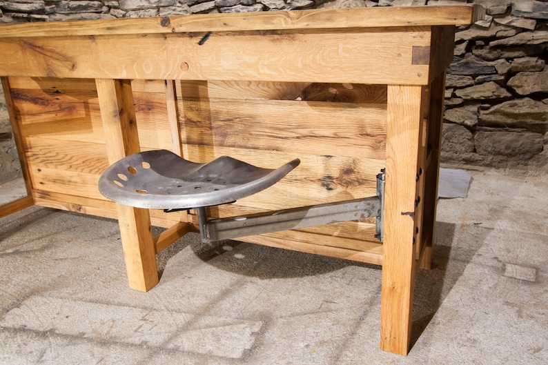 May include: A rustic wooden table with a metal tractor seat attached to the side. The table is made of reclaimed wood and has a natural finish. The seat is made of metal and has a black finish.