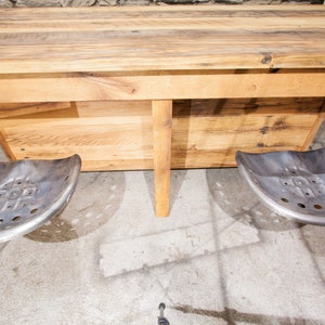 May include: A rustic wooden table with a natural wood finish and two metal tractor seats attached to the underside. The table has a simple design with a rectangular top and a shelf underneath.