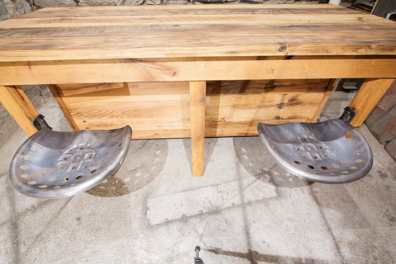 May include: A rustic wooden table with a natural wood finish and two metal tractor seats attached to the underside. The table has a simple design with a rectangular top and a shelf underneath.