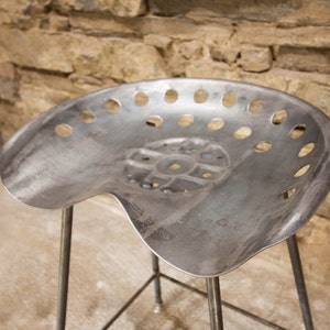 May include: A metal stool with a unique, perforated seat. The seat is shaped like a tractor seat and has a distressed, industrial look.