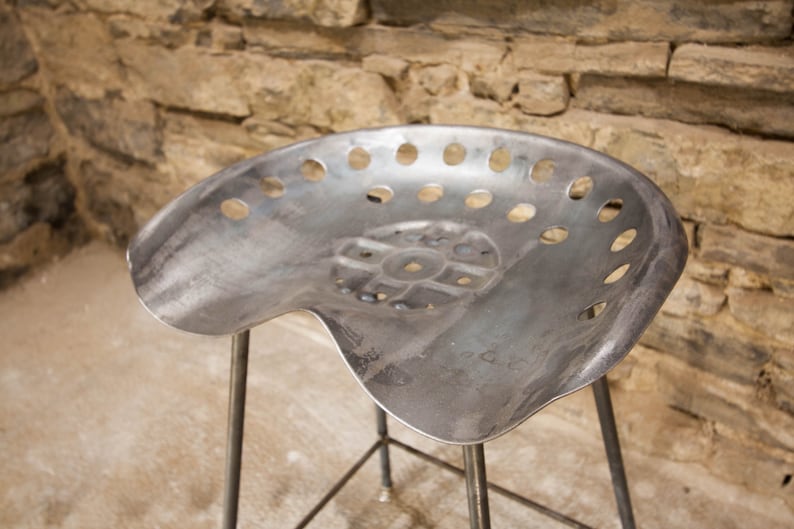 May include: A metal stool with a unique, perforated seat. The seat is shaped like a tractor seat and has a distressed, industrial look.