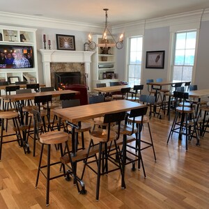 May include: A room featuring several high tables and stools. The tables have light-coloured wooden tops and black metal pipe legs. The stools have wooden seats and black metal frames. The room includes a fireplace, a TV, and large windows.
