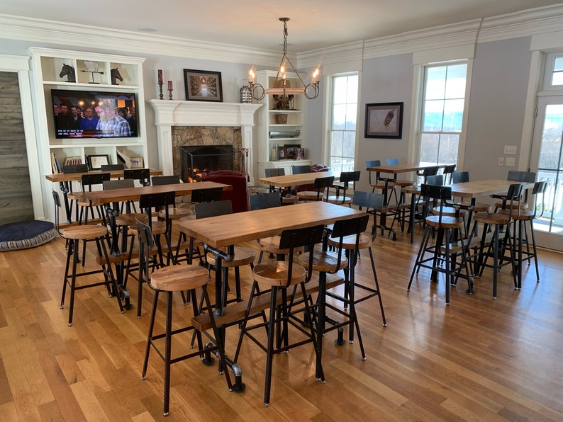 May include: A room featuring several high tables and stools. The tables have light-coloured wooden tops and black metal pipe legs. The stools have wooden seats and black metal frames. The room includes a fireplace, a TV, and large windows.