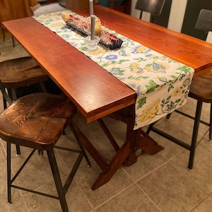 Op de afbeelding: Een houten eettafel met een bloemige tafelrunner en vier zwarte metalen stoelen met houten zittingen. De tafel heeft een rustieke, landelijke stijl.